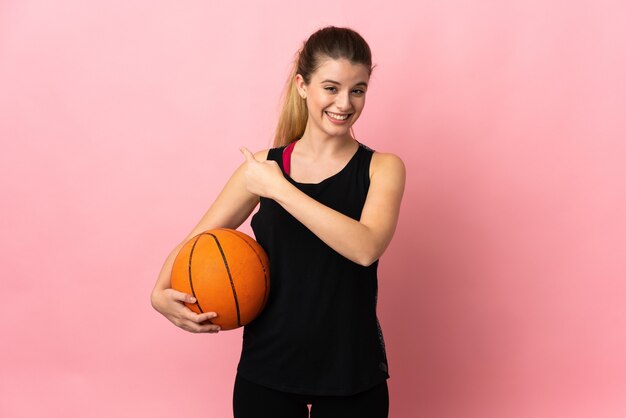 Młoda blond kobieta gra w koszykówkę na białym tle na różowej ścianie, wskazując wstecz