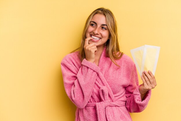 Młoda blond kobieta kaukaski nosi szlafrok, trzymając opaski do depilacji na żółtym tle zrelaksowany, myśląc o czymś, patrząc na miejsce na kopię.