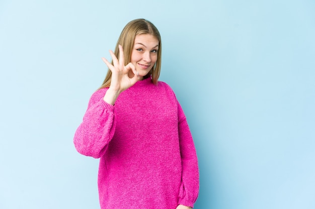 Młoda blond kobieta na białym tle na niebieskiej ścianie, wesoły i pewny siebie, pokazując ok gest.