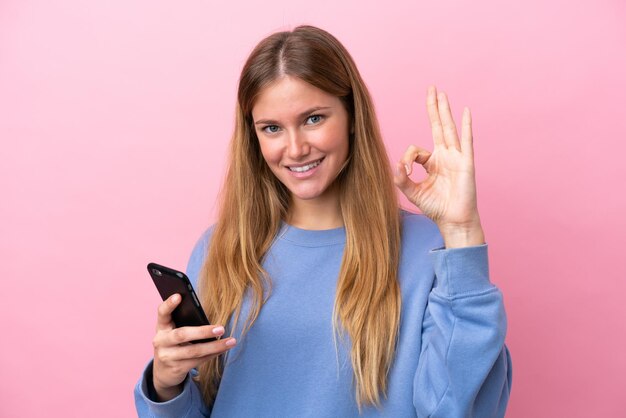 Młoda blond kobieta na białym tle na różowym tle za pomocą telefonu komórkowego i robi znak OK
