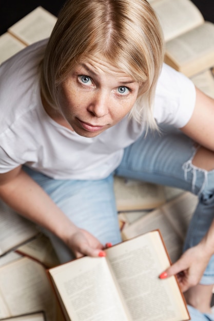 Zdjęcie młoda blond kobieta siedzi na stosie książek i czyta. edukacja, wiedza i hobby. pionowy.