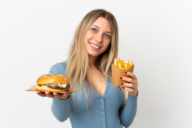 Młoda blond kobieta trzyma smażone frytki i cheeseburgera na białym tle