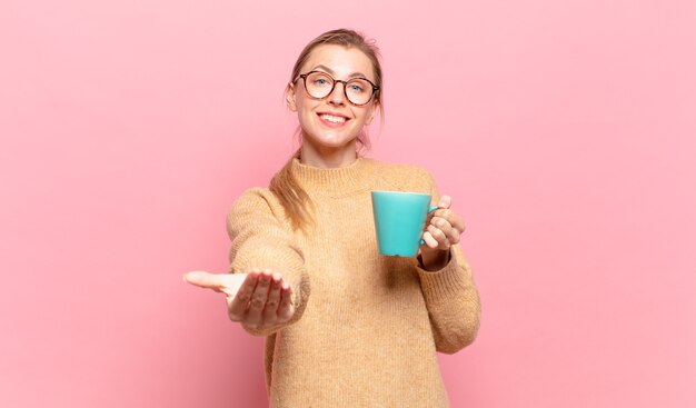 Młoda blond kobieta uśmiecha się radośnie z przyjaznym, pewnym siebie, pozytywnym spojrzeniem, oferując i pokazując przedmiot lub koncepcję. koncepcja kawy