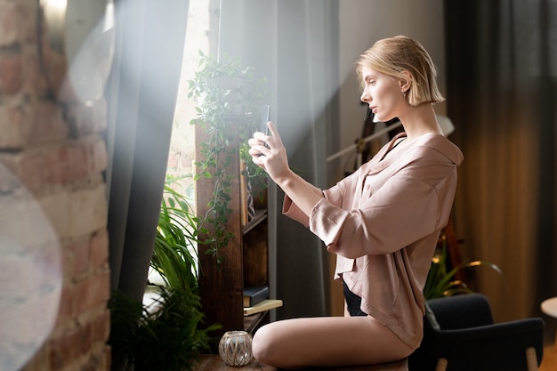 Młoda blond kobieta w czarnej bieliźnie i beżowej jedwabnej piżamie stojąca przed dużym oknem w sypialni i robiąca rano selfie