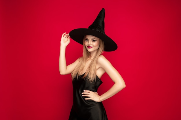 Młoda blond kobieta w czarnym kapeluszu i kostiumu na czerwonym tle. Atrakcyjne kaukaski modelki pozowanie. Halloween, czarny piątek, cyber poniedziałek, sprzedaż, koncepcja jesień. Miejsce. Pewny siebie, uśmiechnięty.