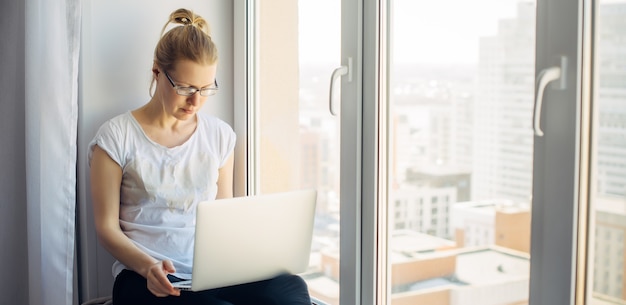 Młoda blond kobieta w okularach w białej koszulce pracuje na laptopie w domu, siedząc na parapecie. Freelancer i biznes zdalny. Tło okna. Skopiuj miejsce.