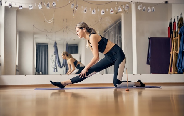 Młoda blond kobieta z idealnym ciałem robi rozciąganie na gumowym dywanie do jogi na siłowni