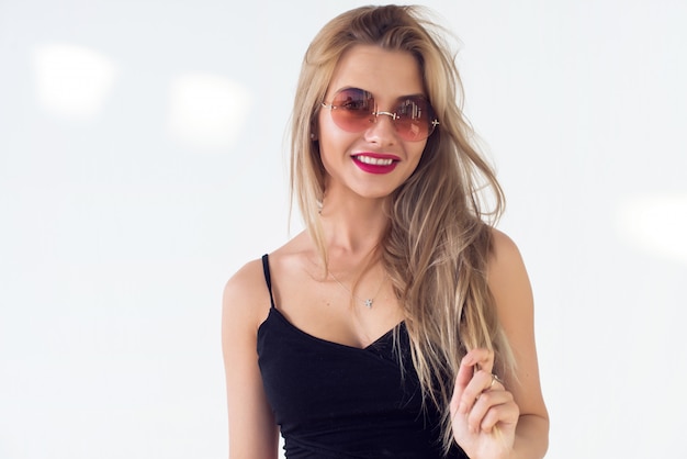 Młoda blond modelka przedstawiająca nowy modny letni wygląd, ubrana w okrągłe okulary przeciwsłoneczne, czerwoną spódnicę i czarny portret z przodu bez rękawów z góry.