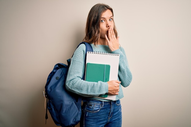 Młoda blond studentka w plecaku i książkach ze szkoły na odizolowanym tle zakrywa usta dłonią zszokowaną wstydem za pomyłkę wyrazem strachu przestraszonego w ciszy tajnej koncepcji