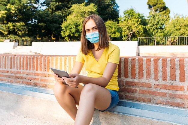 Młoda blond włosy dziewczyna ubrana w żółtą koszulkę, maskę chirurgiczną, patrząc na jej tablet. Koncepcja technologii, stylu życia, relaksu, kwarantanny, covid-19 i omicron