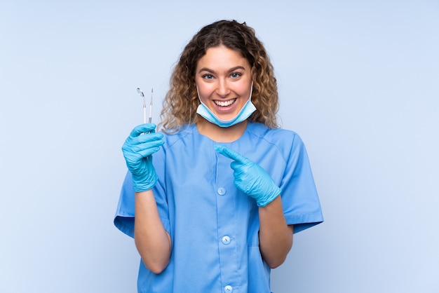 Młoda blondynka dentysta kobieta trzymając narzędzia na białym tle na niebieskiej ścianie palcem wskazującym z boku