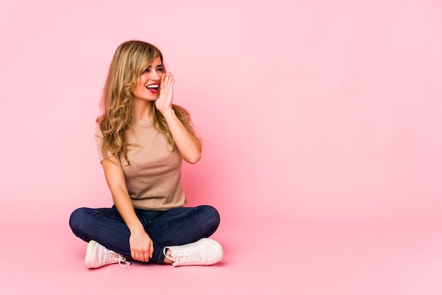 Młoda blondynka kaukaski kobieta siedzi na różowym studio krzycząc i trzymając dłoń w pobliżu otwartych ust.
