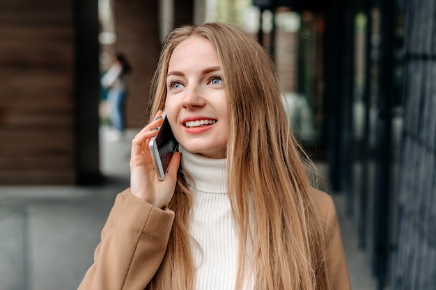 Młoda blondynka Kaukaski kobieta w beżowym płaszczu rozmawia przez telefon komórkowy, uśmiechając się, trzymając folder i patrząc od budynku biurowego. Pracownik korporacyjny. Informatyk