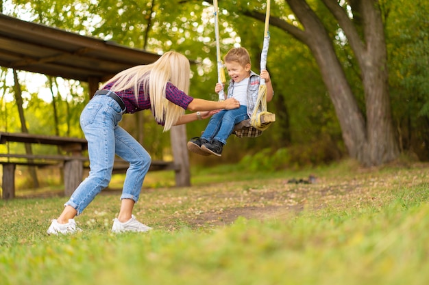 Młoda blondynka mama potrząsa swoim małym synkiem na huśtawce w zielonym parku. Szczęśliwe dzieciństwo.