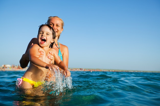 Młoda blondynka matka w niebieskim bikini stojąc w wodzie i bawiąc się z jej małą uśmiechniętą córką