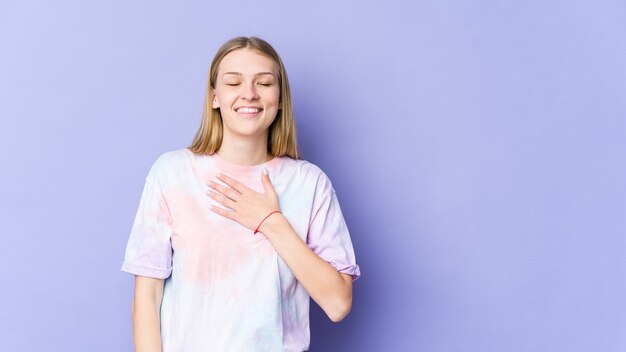 Młoda blondynka na fioletowej ścianie śmieje się głośno, trzymając rękę na piersi.