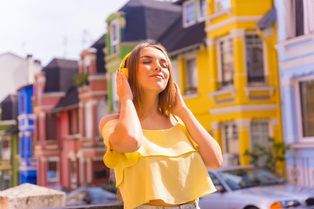 Młoda blondynka na ulicy z domami z kolorowymi fasadami za słuchaniem muzyki online