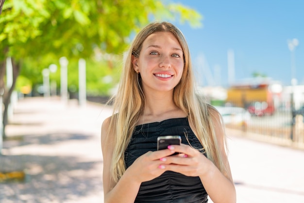 Młoda blondynka na zewnątrz wysyłająca wiadomość za pomocą telefonu komórkowego