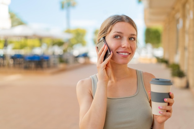 Młoda blondynka na zewnątrz za pomocą telefonu komórkowego i trzymając kawę