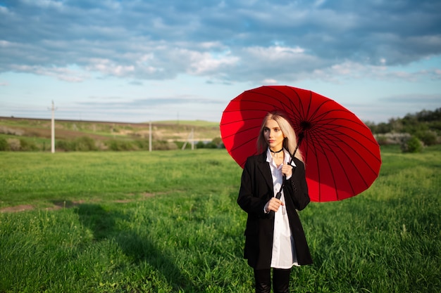 Młoda blondynka piękna dziewczyna, trzymając czerwony parasol, ubrany w czarno-biały garnitur. Tło zachmurzonego nieba i zielonego pola. Kobieta w parku na świeżym powietrzu.