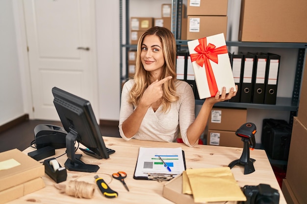 Młoda blondynka pracująca w małym biznesie e-commerce trzymająca prezent uśmiechający się szczęśliwy, wskazujący ręką i palcem