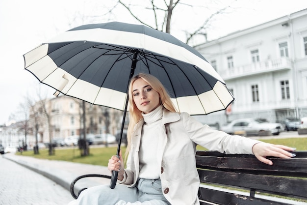 Młoda blondynka siedzi na ławce z parasolem w mieście