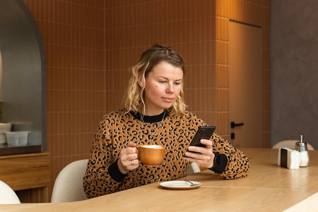 Młoda blondynka siedzi w kawiarni w odcieniach beżu, pije kawę, przewija przez telefon i uśmiecha się