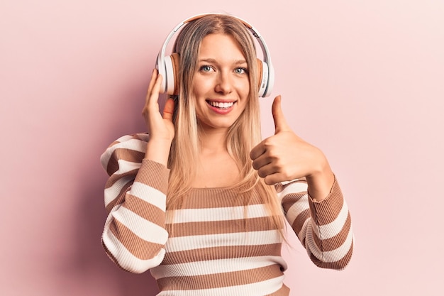Młoda blondynka słucha muzyki za pomocą słuchawek, uśmiechając się radośnie i pozytywnie, kciuk w górę robi doskonały i znak zatwierdzenia
