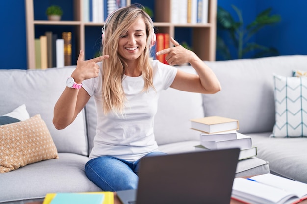 Młoda blondynka studiująca w domu za pomocą laptopa komputerowego uśmiechnięta wesoło pokazująca i wskazująca palcami zęby i usta. koncepcja zdrowia zębów.