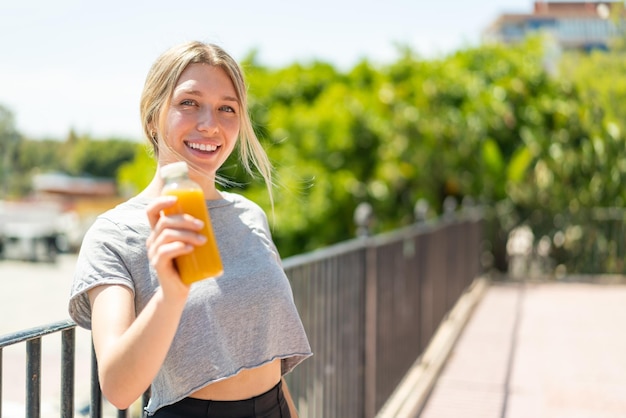 Młoda blondynka trzyma sok pomarańczowy na zewnątrz z radosnym wyrazem twarzy