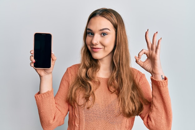 Młoda blondynka trzymająca smartfon pokazujący ekran robi znak ok palcami, uśmiechając się przyjaźnie gestykulując doskonały symbol