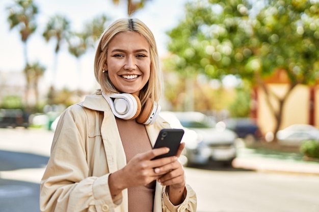 Młoda blondynka uśmiechnięta szczęśliwa za pomocą smartfona i słuchawek w mieście.