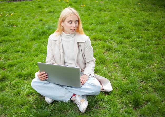 Młoda blondynka używa laptopa siedząc na trawie w parku