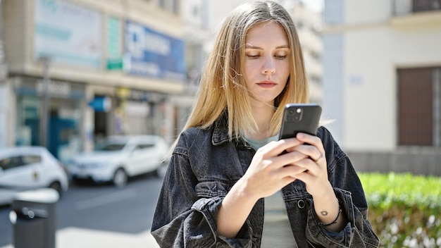 Młoda blondynka używająca smartfona z poważną twarzą na ulicy