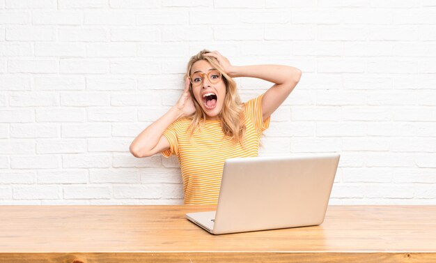 Zdjęcie młoda blondynka w laptopie podnosząc ręce do głowy czuje się szczęśliwy