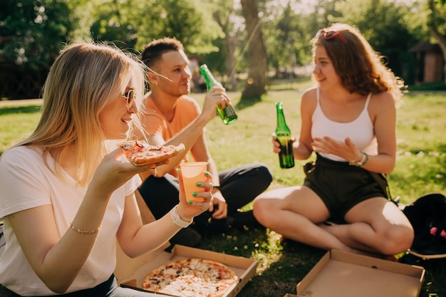 Młoda blondynka w okularach przeciwsłonecznych, picie alkoholu i jedzenie pizzy z przyjaciółmi na świeżym powietrzu.