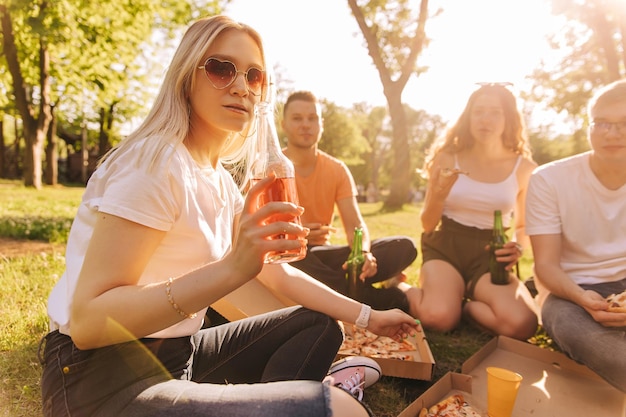 Młoda blondynka w okularach przeciwsłonecznych, picie alkoholu i jedzenie pizzy z przyjaciółmi na świeżym powietrzu.