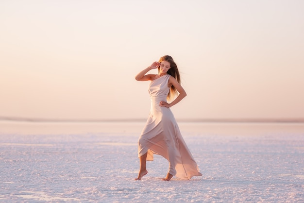 Młoda blondynka w wieczorowej zwiewnej pastelowo różowej, pudrowej sukience stoi boso na białej skrystalizowanej soli. Wycieczka po soli, spacer po wodzie o zachodzie słońca