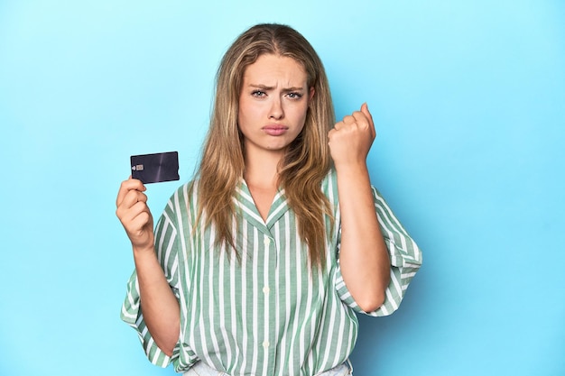 Młoda blondynka z kartą kredytową w niebieskim studiu pokazująca pięść przed kamerą, agresywny wyraz twarzy