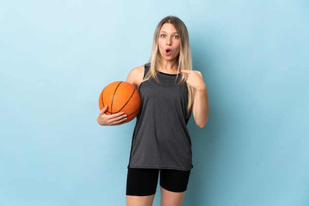 Młoda blondynki kobieta bawić się koszykówkę odizolowywającą na błękit ścianie z niespodzianka wyrazem twarzy