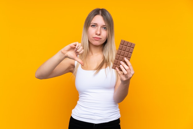Młoda blondynki kobieta nad odosobnioną błękit ścianą bierze czekoladową pastylkę robi złemu sygnałowi