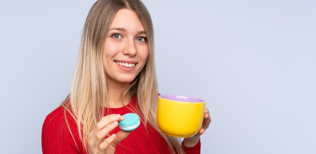 Młoda blondynki kobieta nad odosobnioną błękit ścianą trzyma kolorowych Francuskich macarons i filiżankę mleka