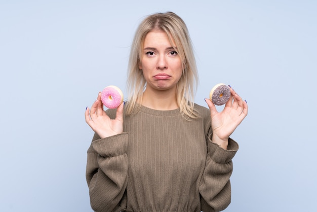 Młoda blondynki kobieta nad odosobnionymi błękit ściany mienia donuts z smutnym wyrażeniem