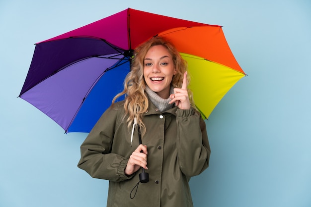 Młoda blondynki kobieta trzyma parasol odizolowywający na błękit ścianie wskazuje w górę doskonałego pomysłu