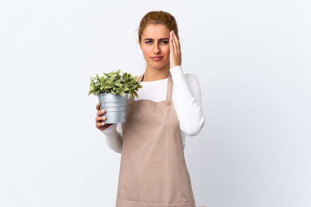 Młoda blondynki ogrodniczki kobiety dziewczyna trzyma rośliny nad odosobnioną biel ścianą nieszczęśliwą i sfrustowaną z czymś. Negatywny wyraz twarzy