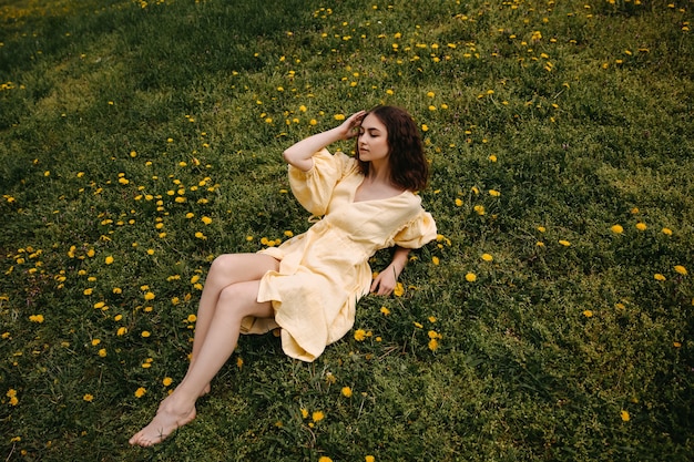 Młoda boso kobieta w żółtej sukience relaksuje się na polu z zieloną trawą i mniszkami