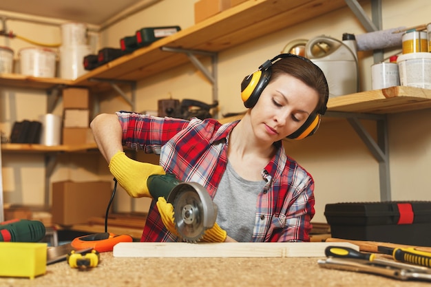 Młoda brązowowłosa kobieta w koszuli w kratę, szara koszulka, słuchawki izolowane akustycznie, żółte rękawiczki pracujące w warsztacie stolarskim przy drewnianym stole z różnymi narzędziami, piłowanie drewna piłą mechaniczną.