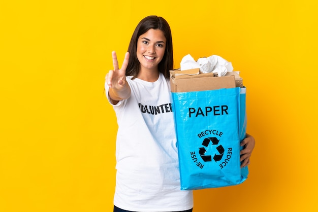 Młoda brazylijska dziewczyna trzyma worek recyklingu pełen papieru do recyklingu na białym tle na żółtej ścianie, uśmiechając się i pokazując znak zwycięstwa