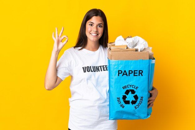 Młoda brazylijska dziewczyna trzyma worek recyklingu pełen papieru do recyklingu na żółtym, pokazując znak ok palcami