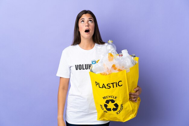 Młoda brazylijska kobieta trzyma torbę pełną plastikowych butelek do recyklingu na białym tle na fioletowy patrząc w górę iz zaskoczonym wyrazem twarzy